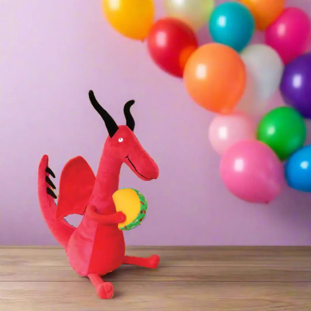 Red dragon plush toy with a yellow ball in front of colorful balloons against a pink wall.