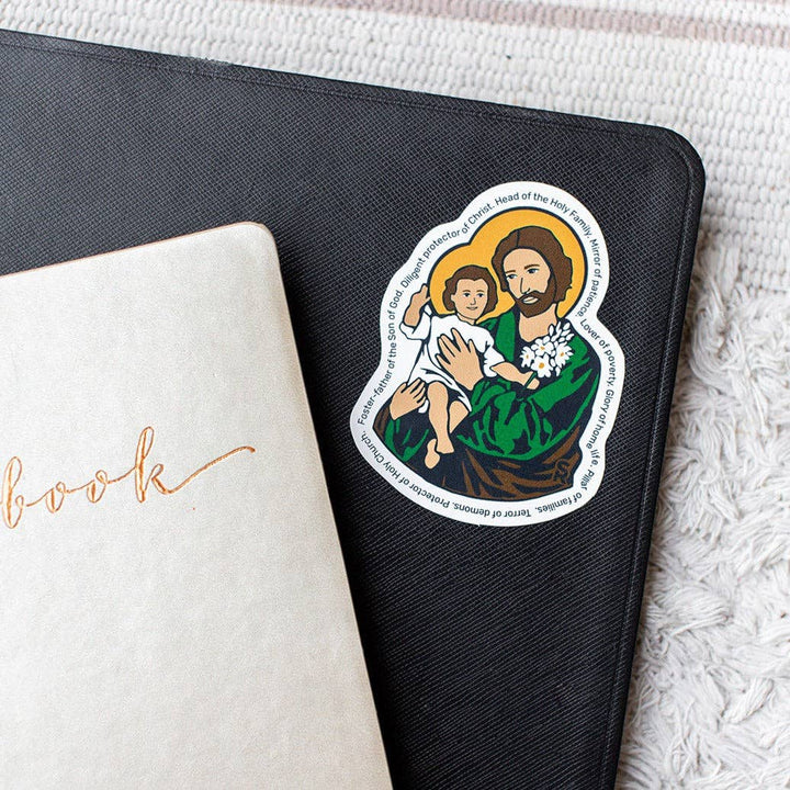 Sticker of a religious figure holding a child on a black notebook with a white book underneath.