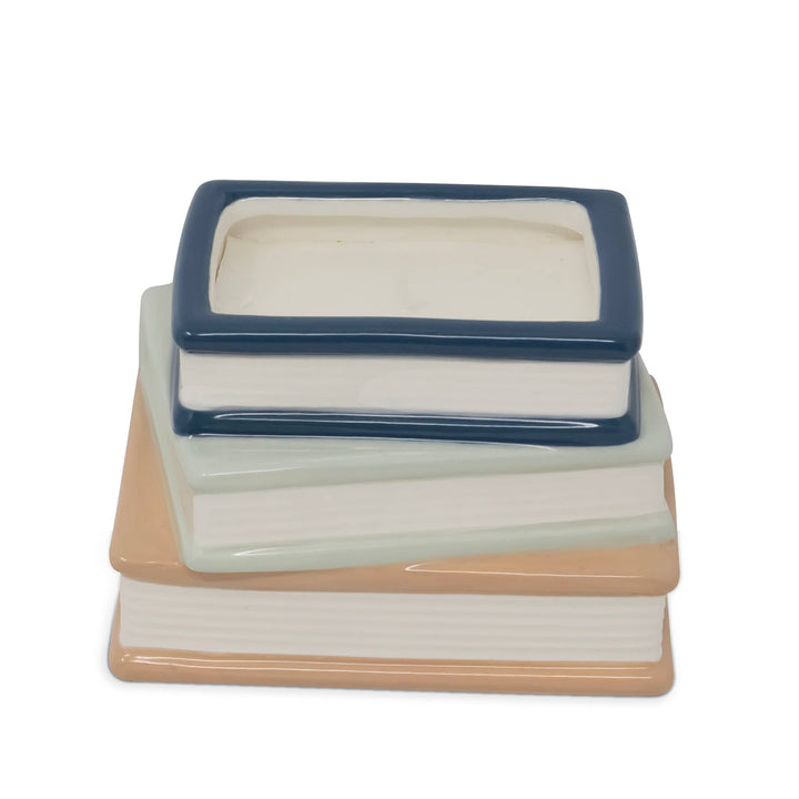 A ceramic stack of three books with a rectangular box lid on top, featuring cream pages, a peach-colored bottom book, a pale mint middle book, and a dark blue top border.