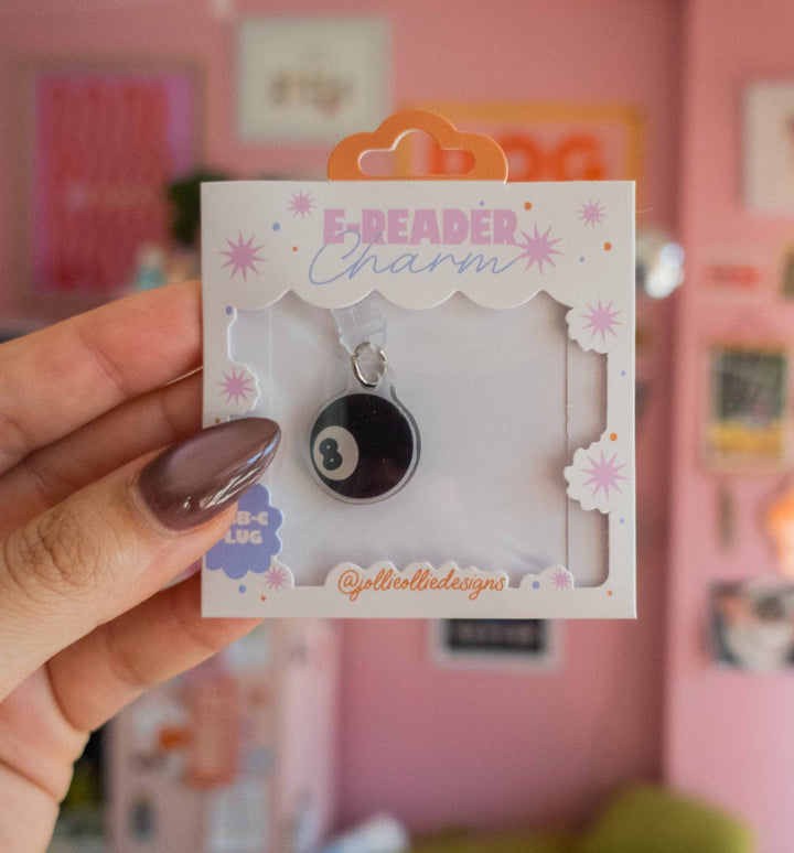 A hand with mauve nail polish holds a small retail card featuring a black-and-white 8-ball charm labeled &