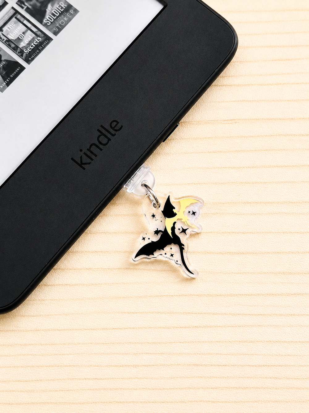 A close-up of a black Kindle e-reader lying on a light wood surface, with a small hanging dragon charm attached near the bottom edge. The charm depicts a black-and-white dragon with a yellow and small star details.