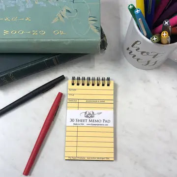 Small yellow notepad with a pen on a white surface, with books and a mug in the background.