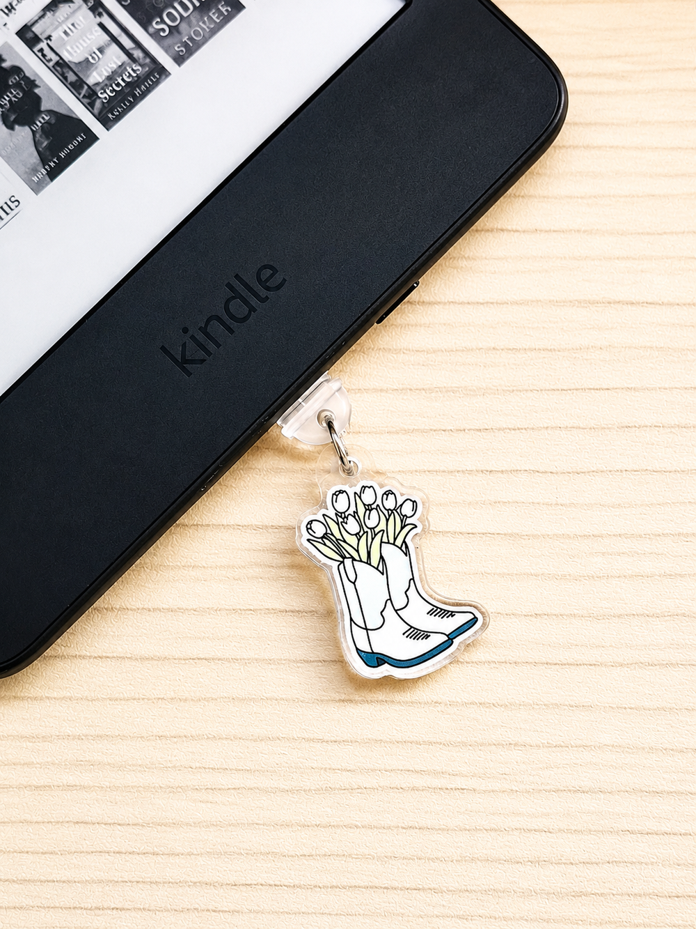 A small enamel charm shaped like white boots filled with tulips hangs from a clear clip on the edge of a black Kindle e-reader, placed on a light wood surface.