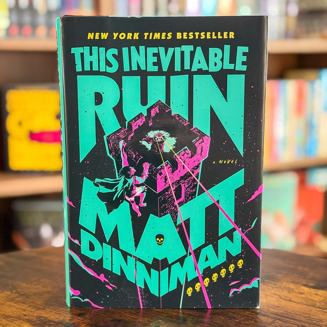 The book cover of 'This Inevitable Ruin' by Matt Dinniman, featuring a black background with bold teal and hot pink typography and a central illustration of a person falling into a dark abyss, displayed on a wooden table with a blurred bookstore background.