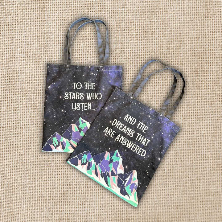 Two tote bags with starry night design and mountain illustrations on a textured beige background.