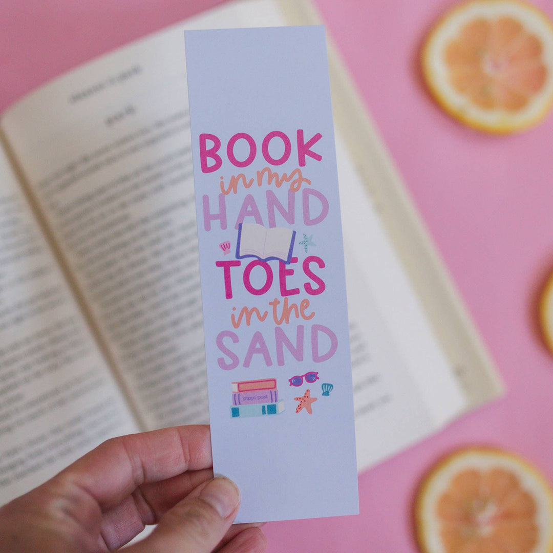 Hand holding a colorful bookmark with text over an open book on a pink background.