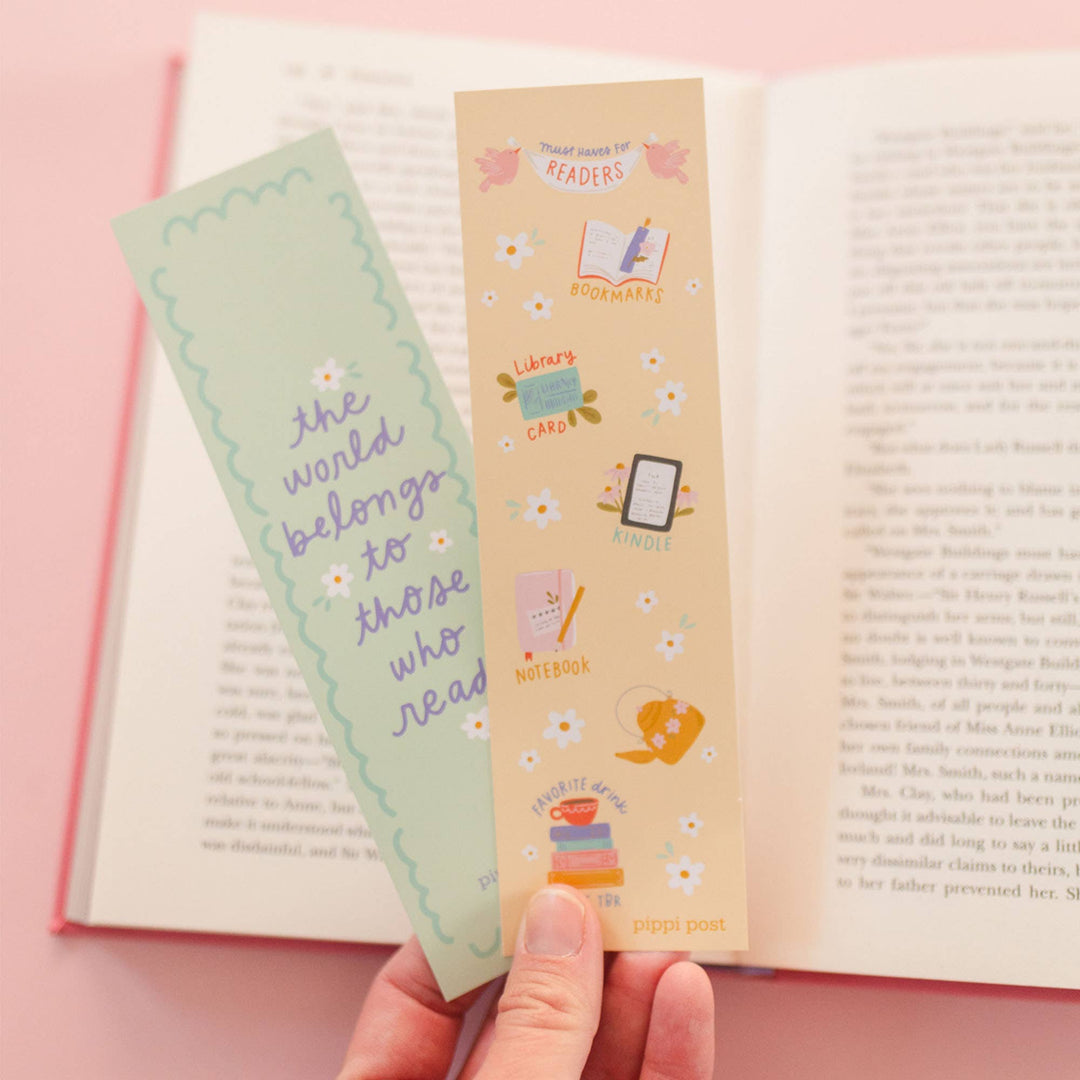 Two bookmarks, one green and one beige with illustrations, held over an open book on a pink background.