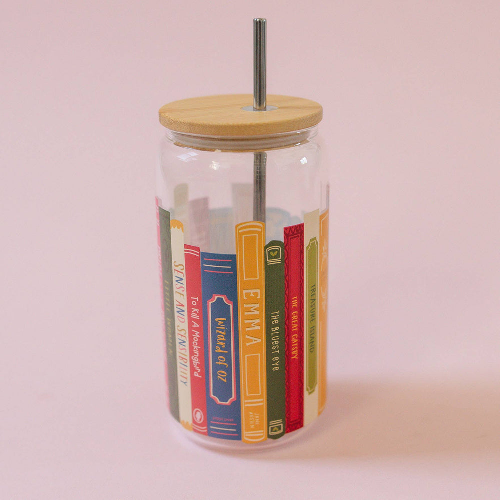 Glass tumbler with wooden lid and metal straw featuring book spines design on a pink background