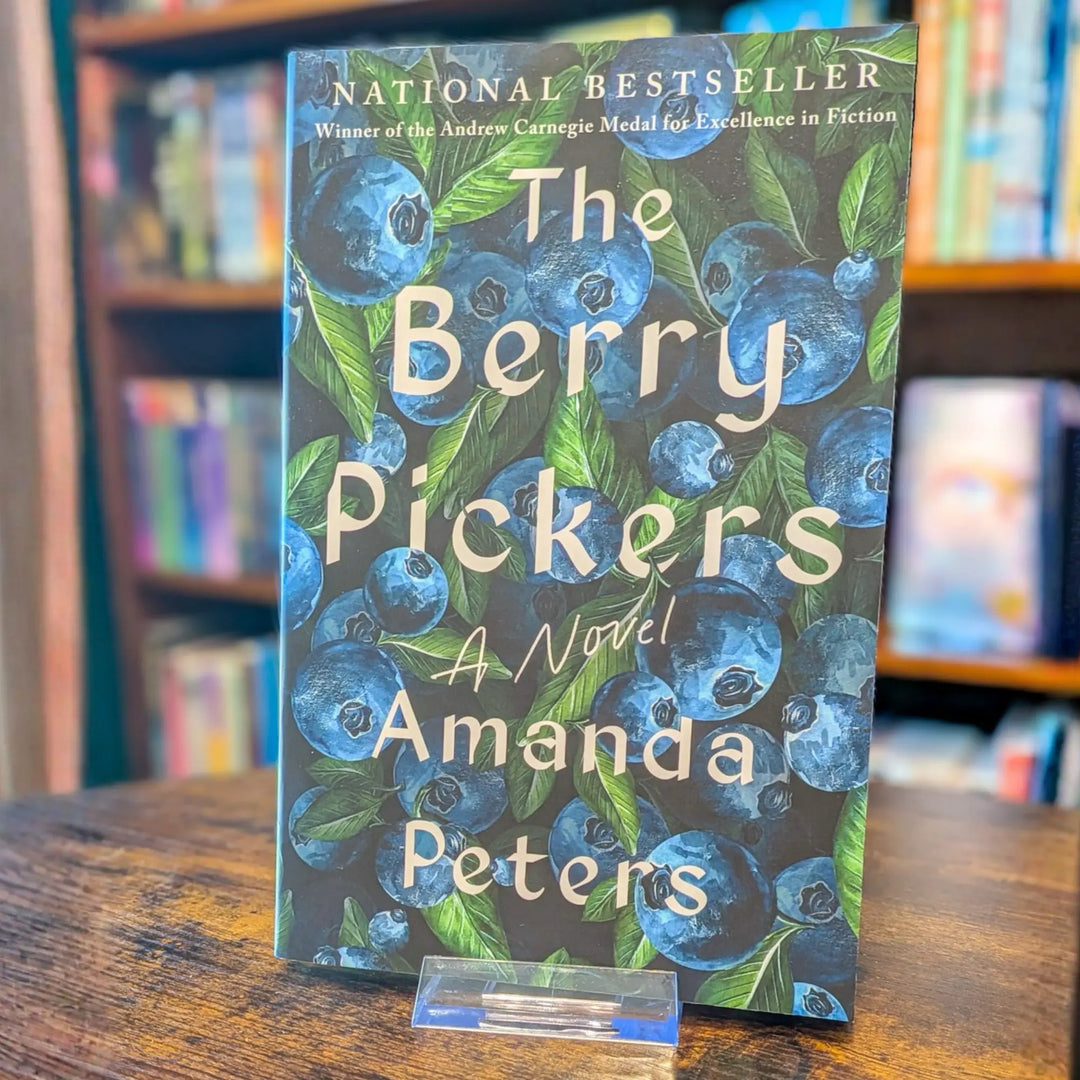The book cover of 'The Berry Pickers' by Amanda Peters, featuring a dark blue and green blueberry pattern, displayed on a wooden table in a blurred library or bookstore setting.