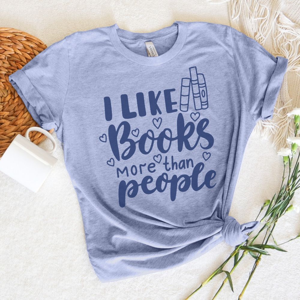 Light blue t-shirt with 'I Like Books More Than People' text on a white surface with wicker basket and flowers.