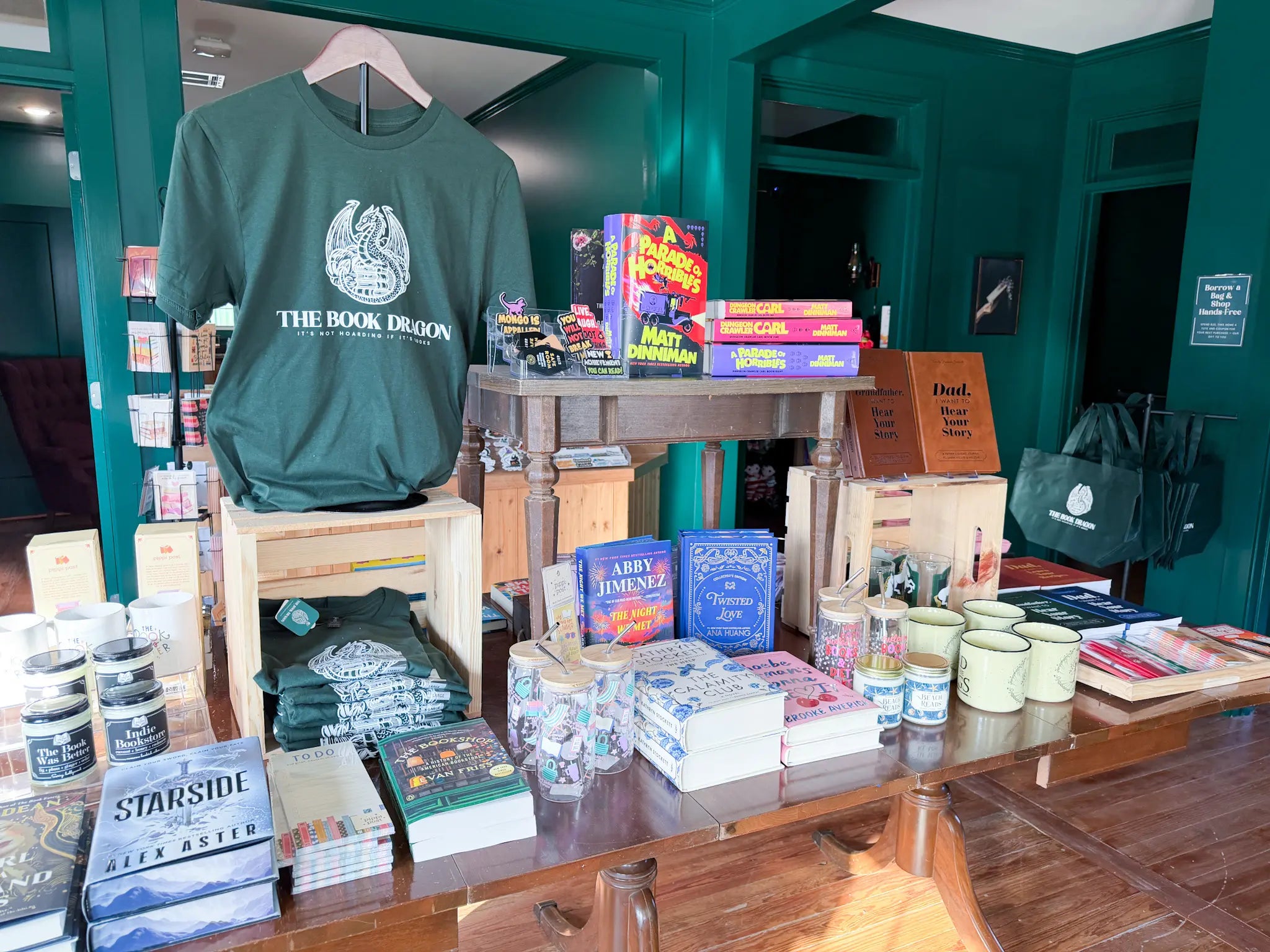 Display of books and merchandise in a store with a green t-shirt labeled 'The Book Dragon' on a wooden table.