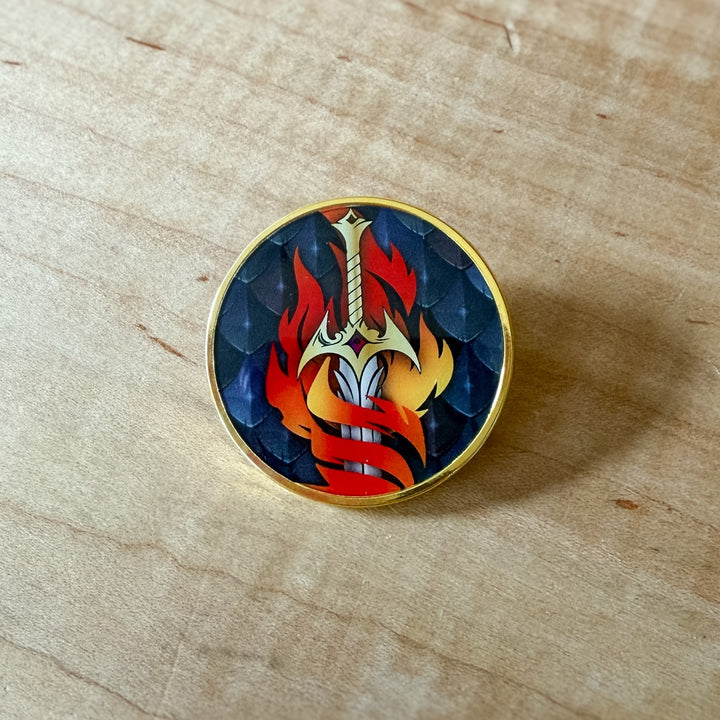 Round enamel pin with sword and flame design on a wooden surface