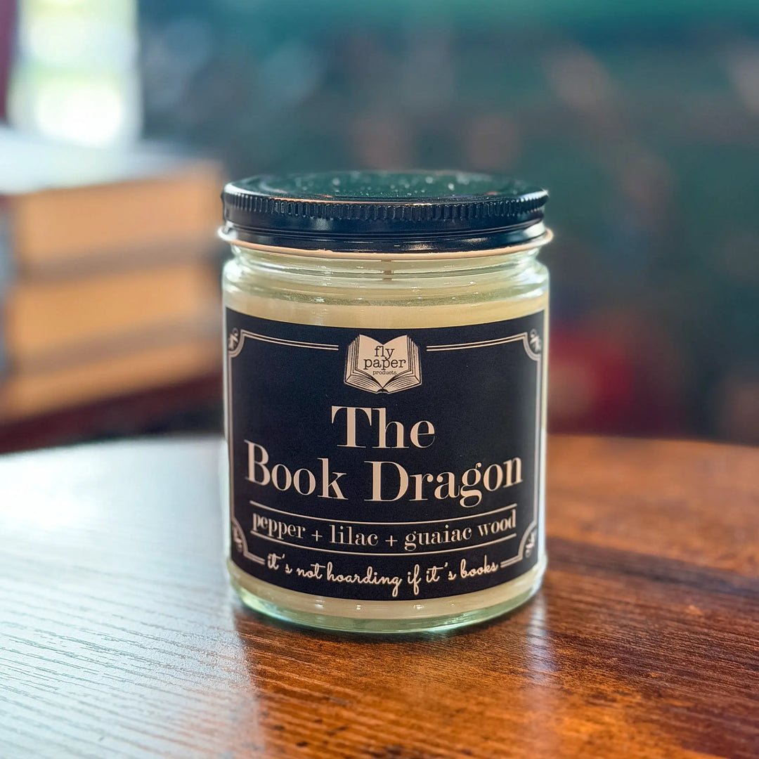 Jar of candle labeled 'The Book Dragon' on a wooden surface with a blurred background