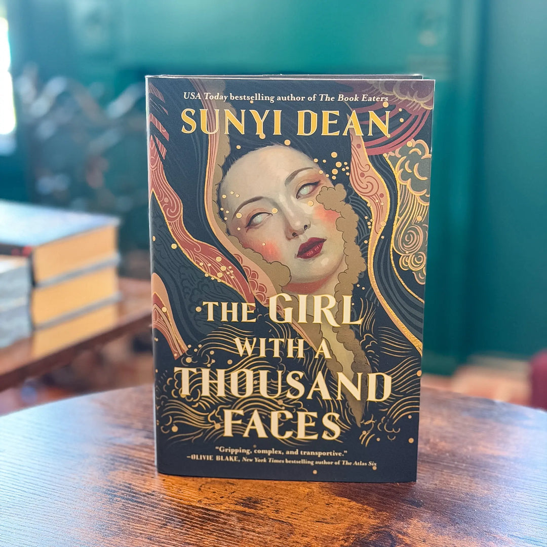 Book titled 'The Girl with a Thousand Faces' by Sunyi Dean on a wooden surface.