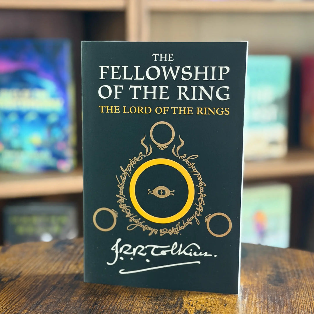 Book titled 'The Fellowship of the Ring' from 'The Lord of the Rings' series on a wooden surface with a blurred bookshelf background.