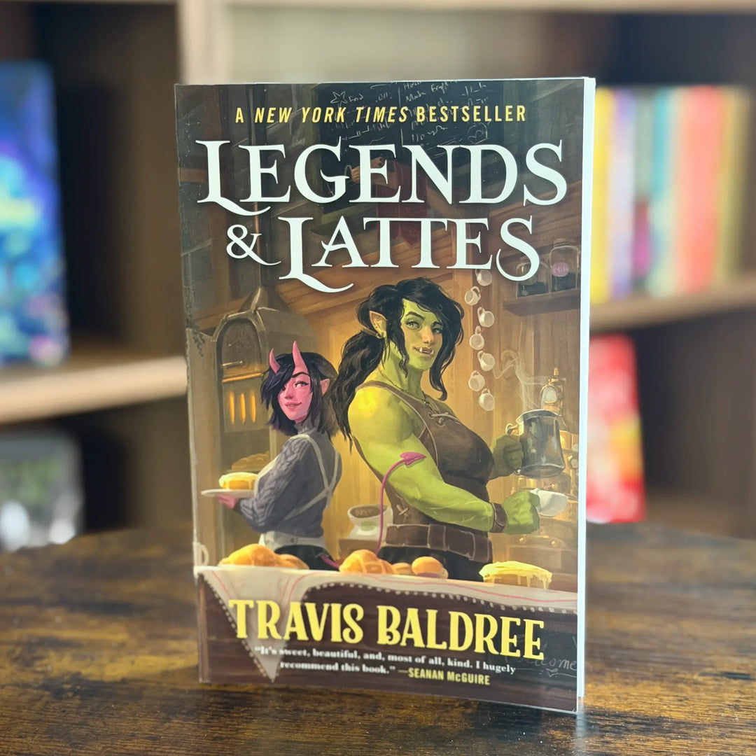 Book titled 'Legends & Lattes' by Travis Baldree on a wooden surface with a blurred bookshelf background.