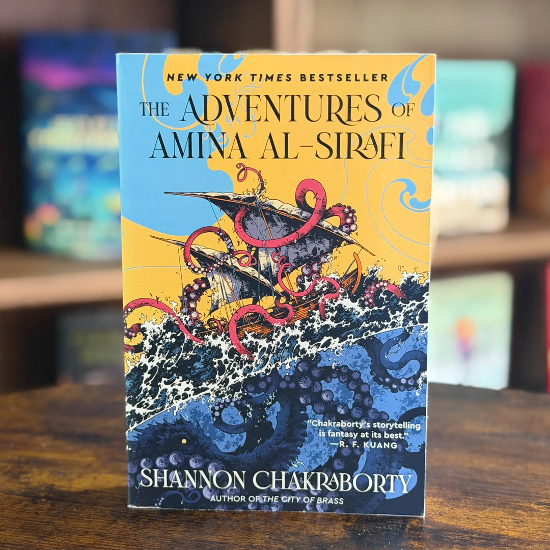 Book cover of 'The Adventures of Amina Al-Sirafi' by Shannon Chakraborty on a wooden surface with blurred books in the background.