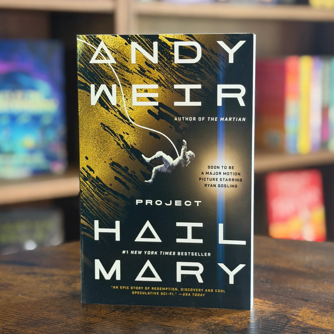 Book cover of 'Project Hail Mary' by Andy Weir on a wooden surface with a blurred bookshelf background.