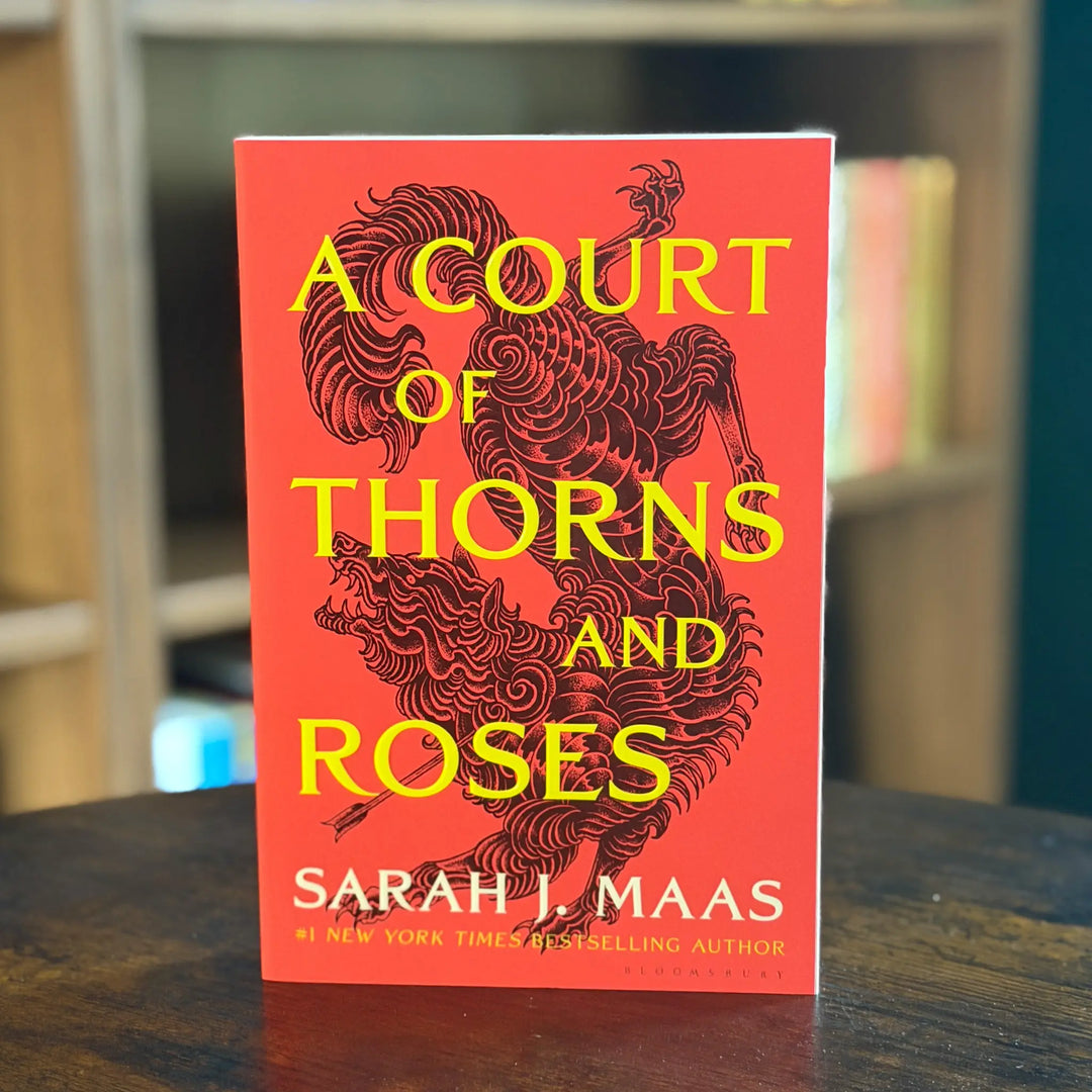 Book titled 'A Court of Thorns and Roses' by Sarah J. Maas on a wooden surface with a blurred bookshelf background.