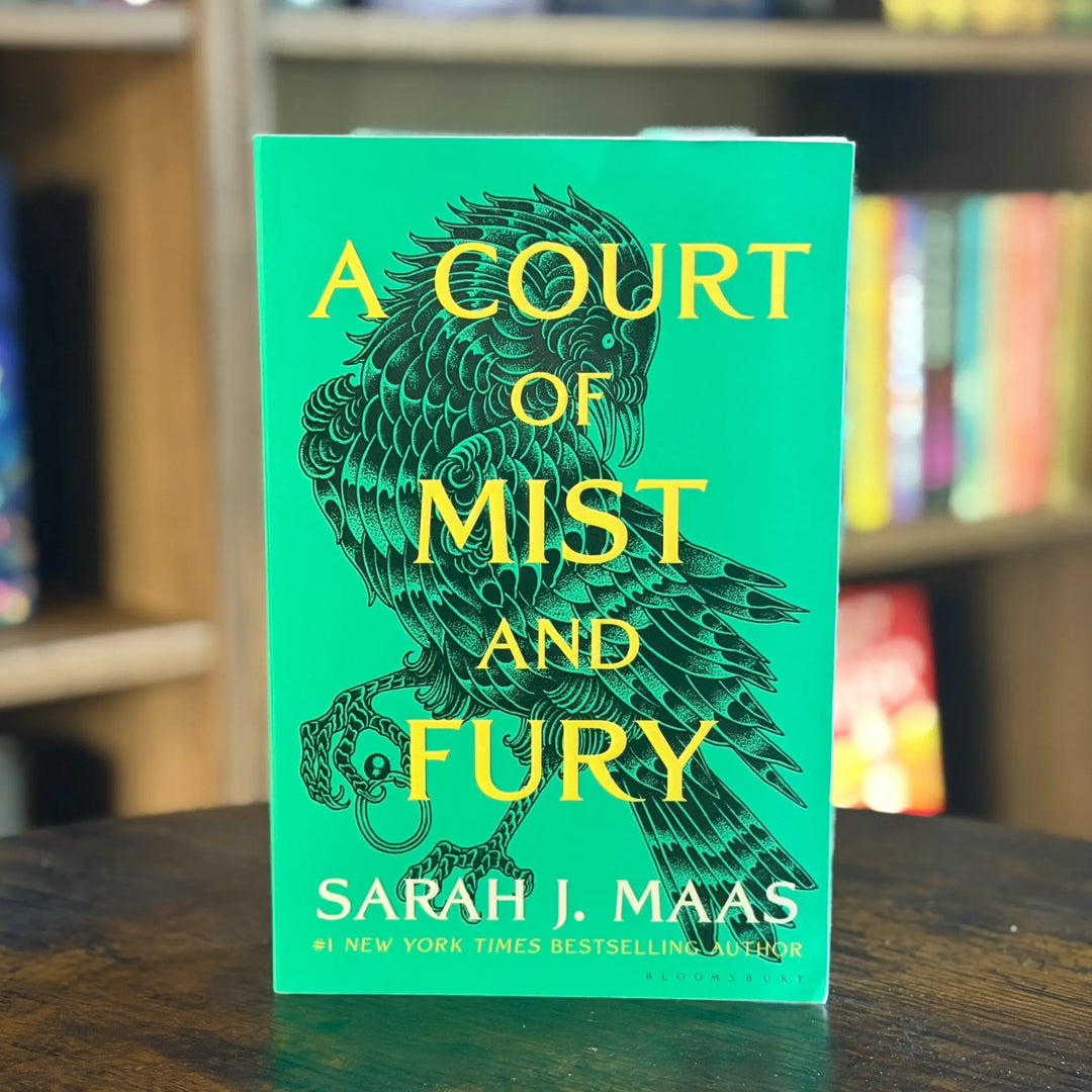 Book titled 'A Court of Mist and Fury' by Sarah J. Maas on a table with a blurred bookshelf background.