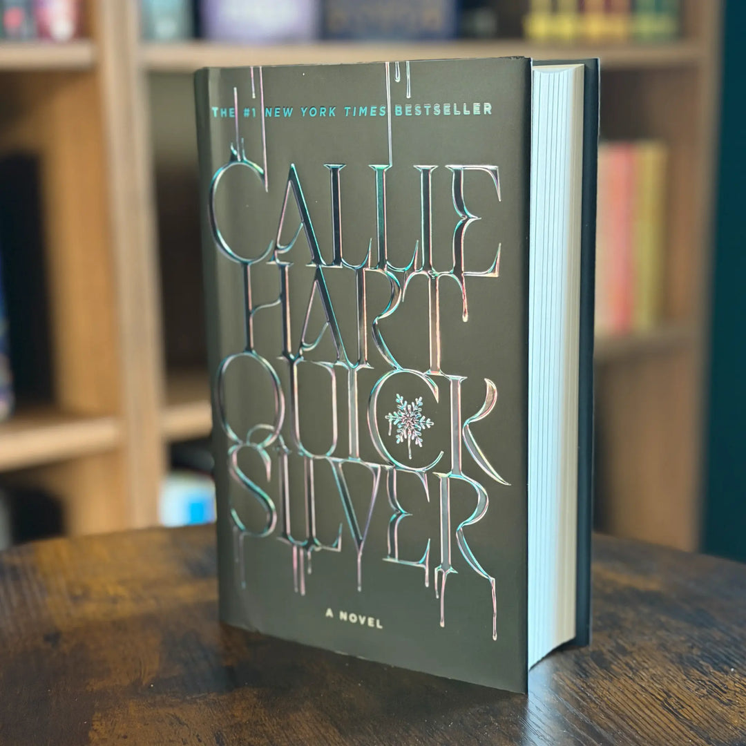 Book titled 'Quick Silver' by Callie Hart on a wooden surface with a blurred bookshelf in the background.