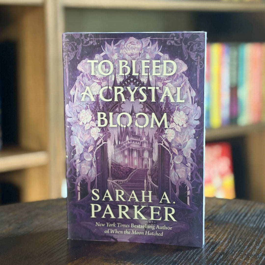 Book titled 'To Bleed a Crystal Bloom' by Sarah A. Parker on a table with a blurred bookshelf background.