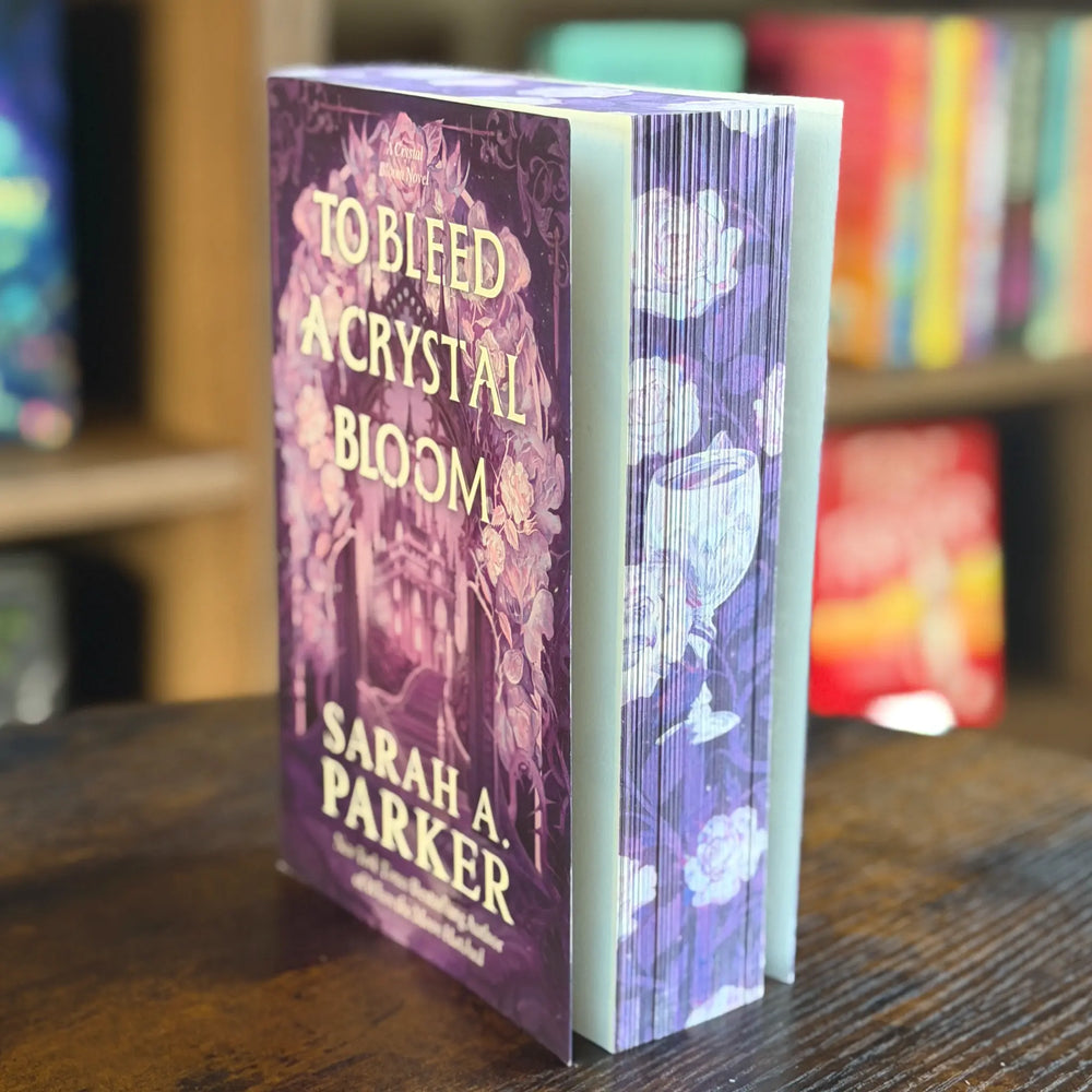 Book titled 'To Bleed a Crystal Bloom' by Sarah A. Parker on a wooden surface with a blurred bookshelf in the background.