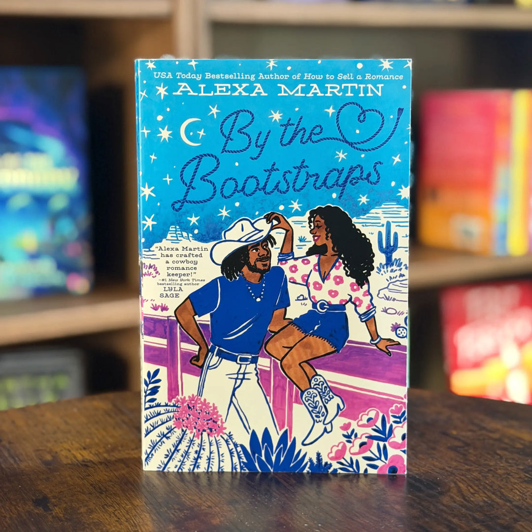 Book titled 'By the Bootstraps' by Alexa Martin on a wooden surface with a blurred bookshelf background.
