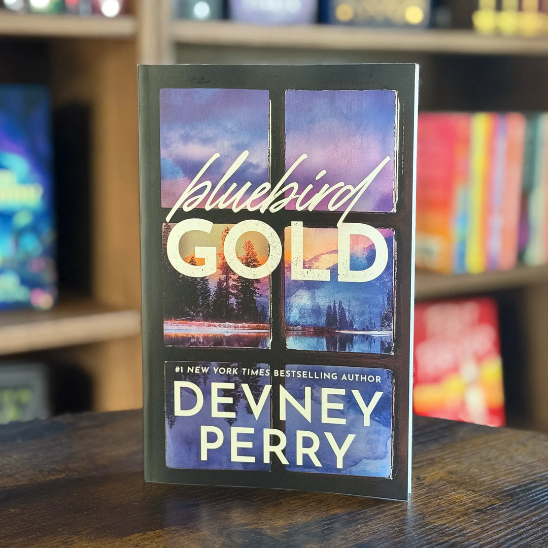 Book titled 'Bluebird Gold' by Devney Perry on a wooden surface with a blurred bookshelf background.