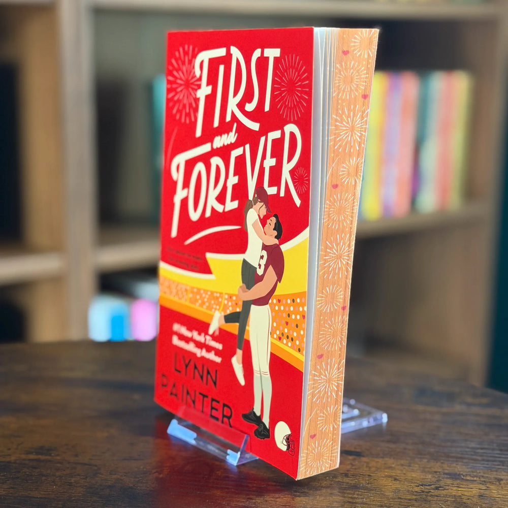 Book titled 'First and Forever' by Lynn Painter on a wooden surface with a blurred bookshelf background.