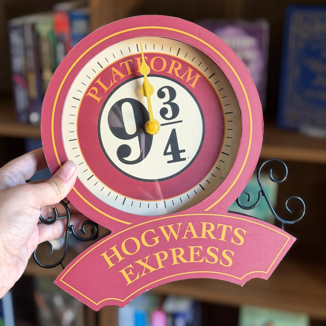 Clock with 'Platform 9 3/4, Hogwarts Express' design held by a hand against a bookshelf background.