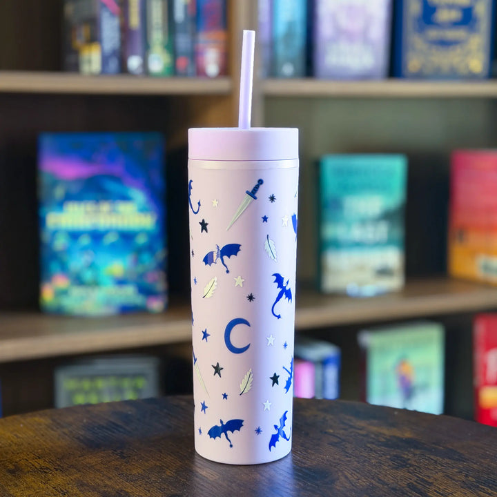 Purple tumbler with blue designs and a straw on a wooden surface with a bookshelf in the background