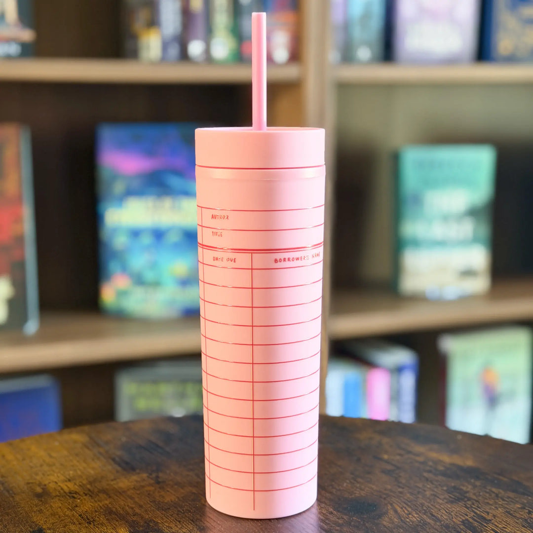 Pink tumbler with a straw on a wooden surface with a bookshelf in the background