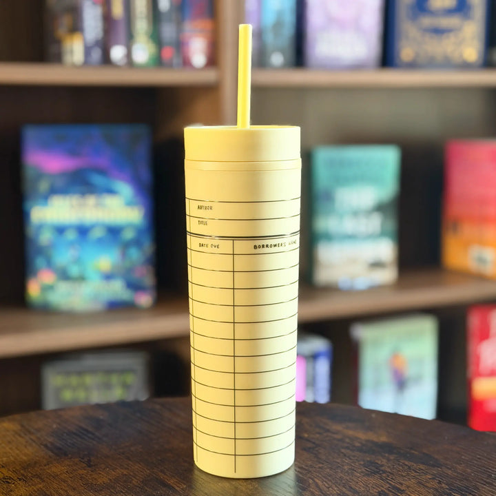 Yellow tumbler with a straw on a wooden surface with a bookshelf in the background