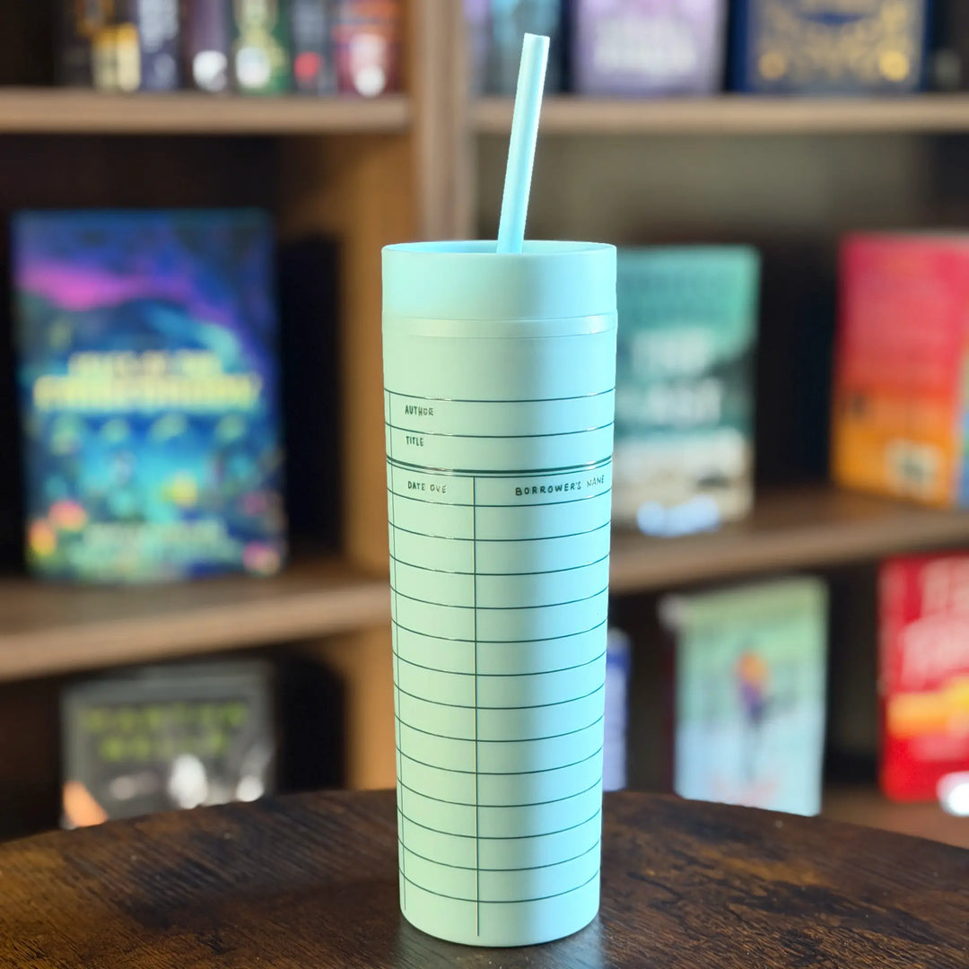 Light blue tumbler with a straw on a wooden surface with a bookshelf in the background
