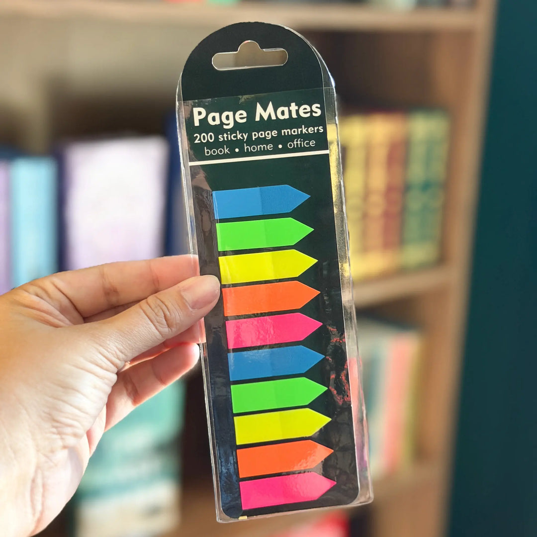 Pack of colorful Page Mates sticky page markers held by a hand with a blurred bookshelf background.