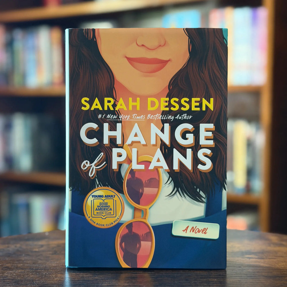 Book titled 'Change of Plans' by Sarah Dessen on a wooden surface with a blurred bookshelf background.