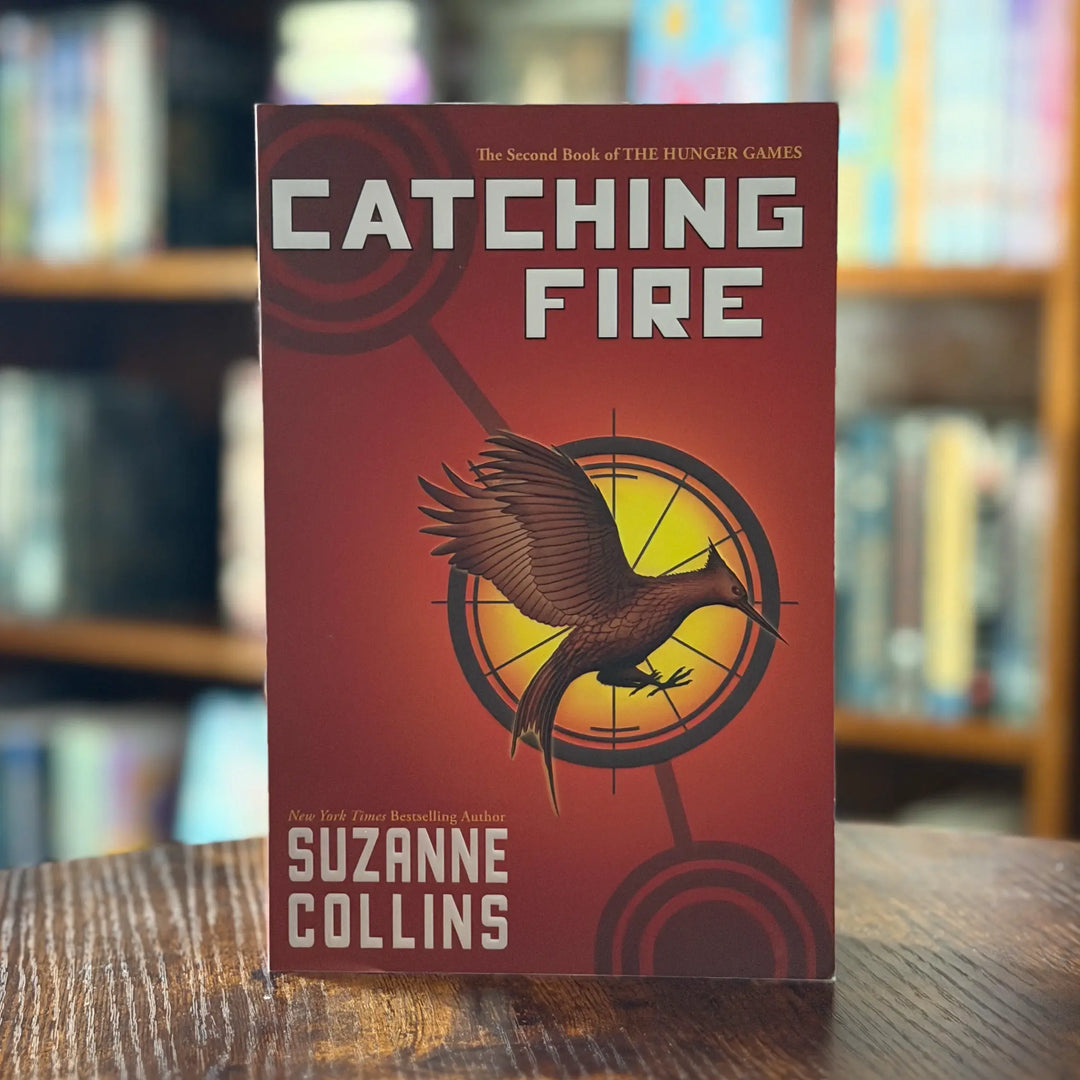 Book titled 'Catching Fire' by Suzanne Collins on a wooden surface with a blurred bookshelf background.