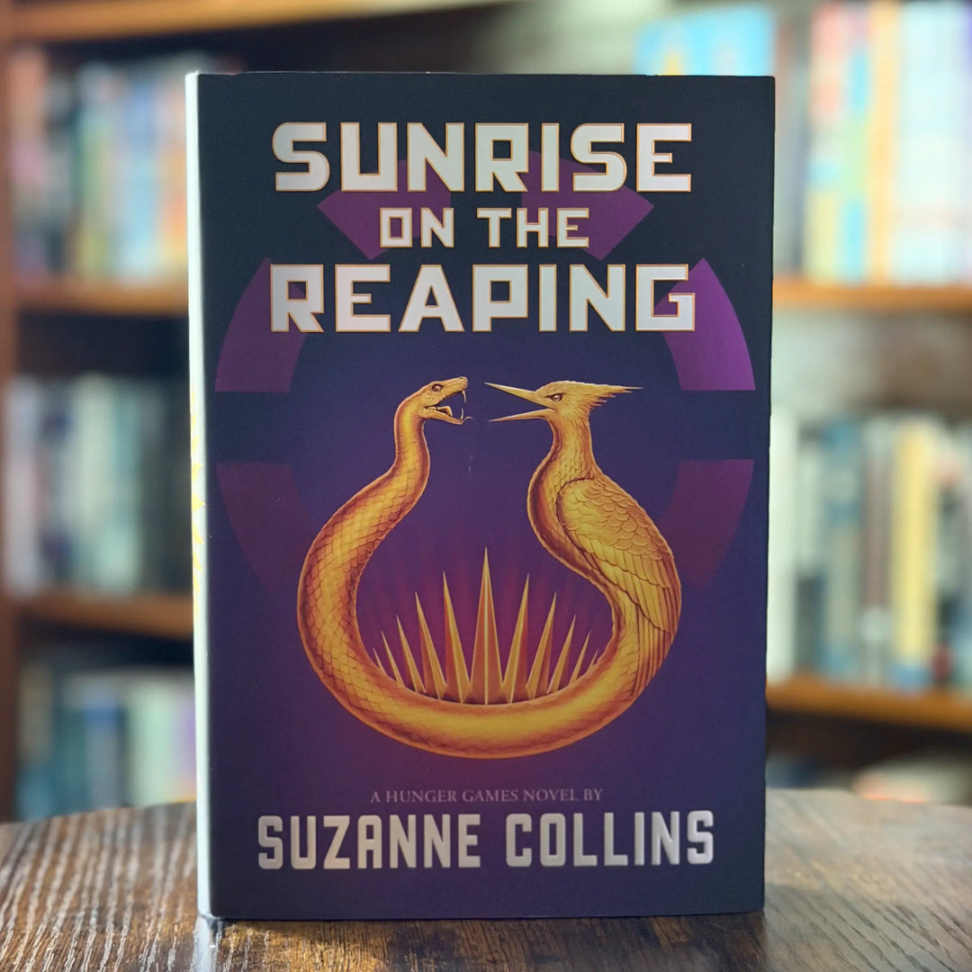 Book titled 'Sunrise on the Reaping' by Suzanne Collins with a purple and gold design on a blurred bookshelf background.