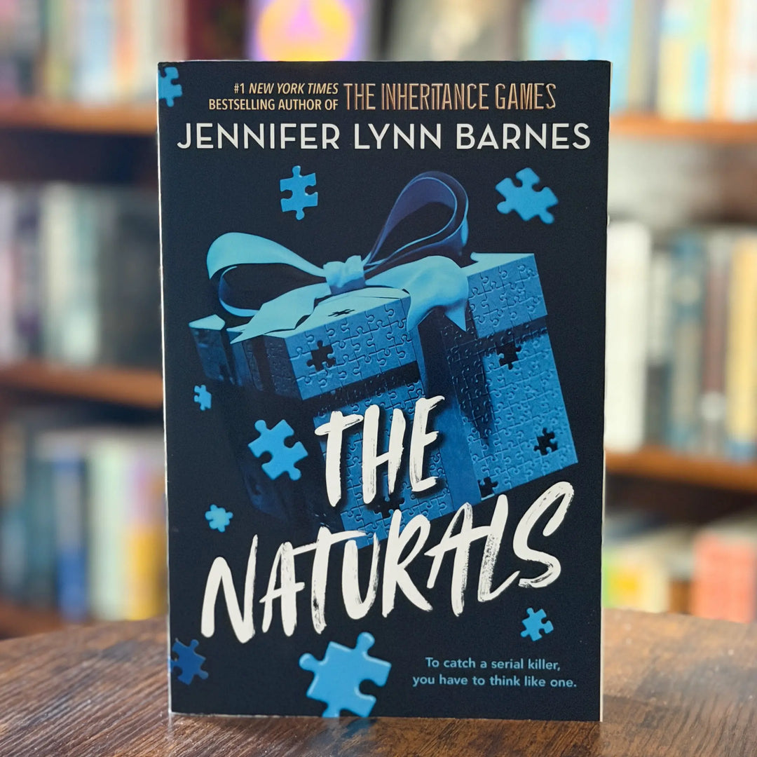 Book cover of 'The Naturals' by Jennifer Lynn Barnes on a wooden surface with a blurred bookshelf background.