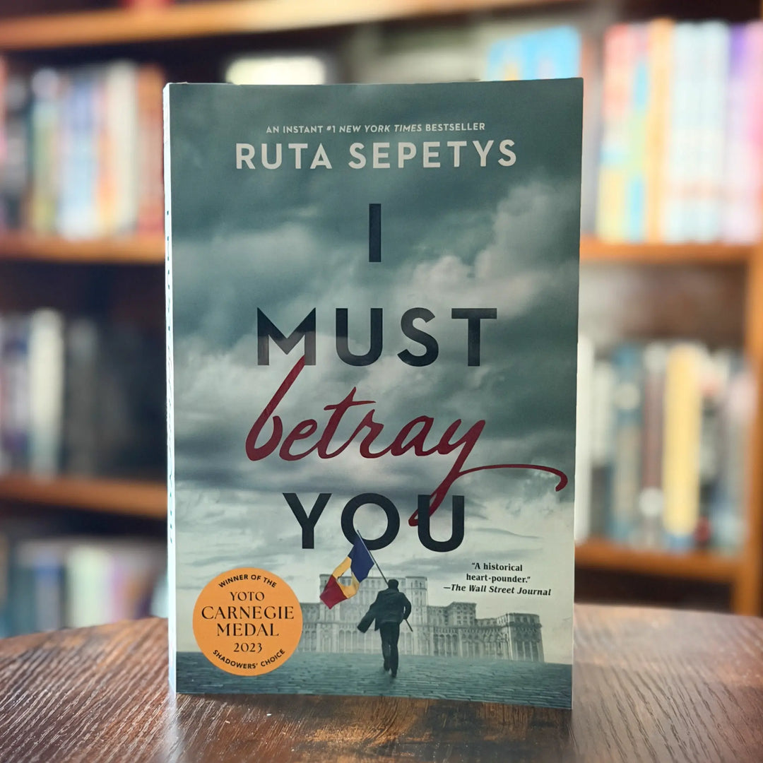 Book titled 'I Must Betray You' by Ruta Sepetys on a wooden surface with a blurred bookshelf background.