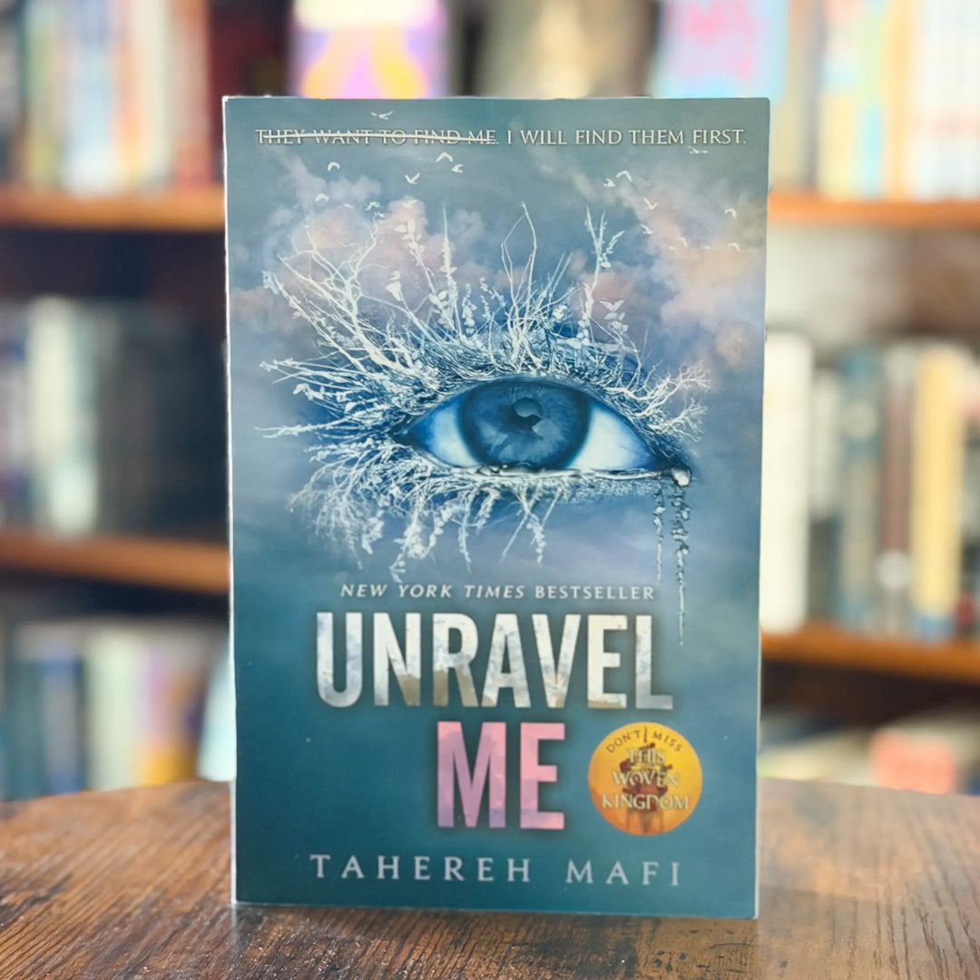 Book titled 'Unravel Me' by Tahereh Mafi on a wooden surface with a blurred bookshelf background.