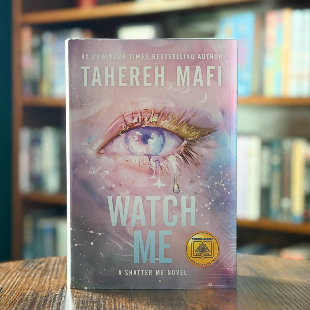 Book titled 'Watch Me' by Tahereh Mafi on a wooden surface with a blurred bookshelf background