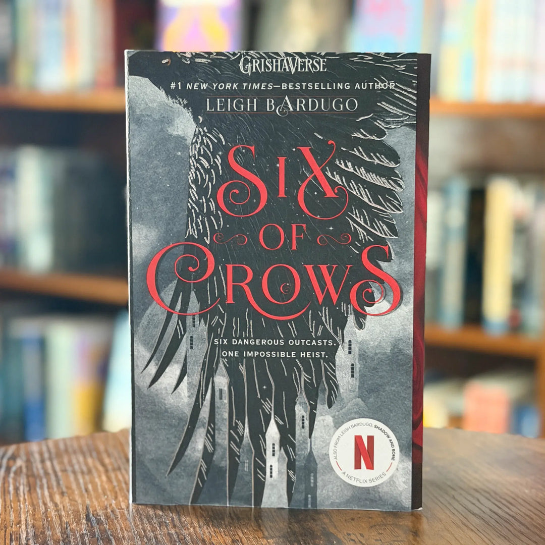 Book titled 'Six of Crows' by Leigh Bardugo with a dark cover featuring crows and a cityscape, placed on a wooden surface with a blurred bookshelf background.