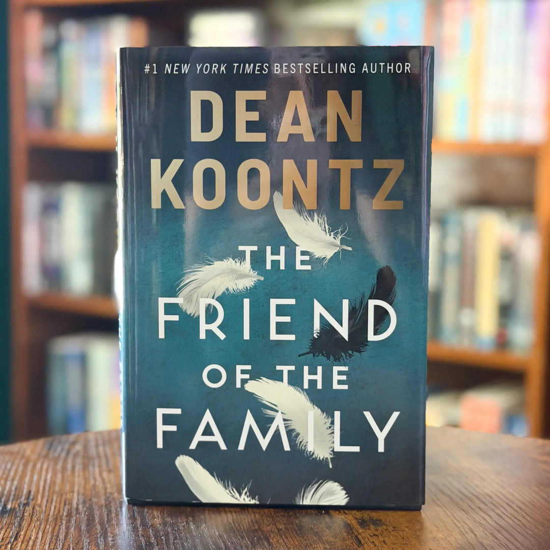 Book titled 'The Friend of the Family' by Dean Koontz on a wooden surface with a blurred bookshelf background.
