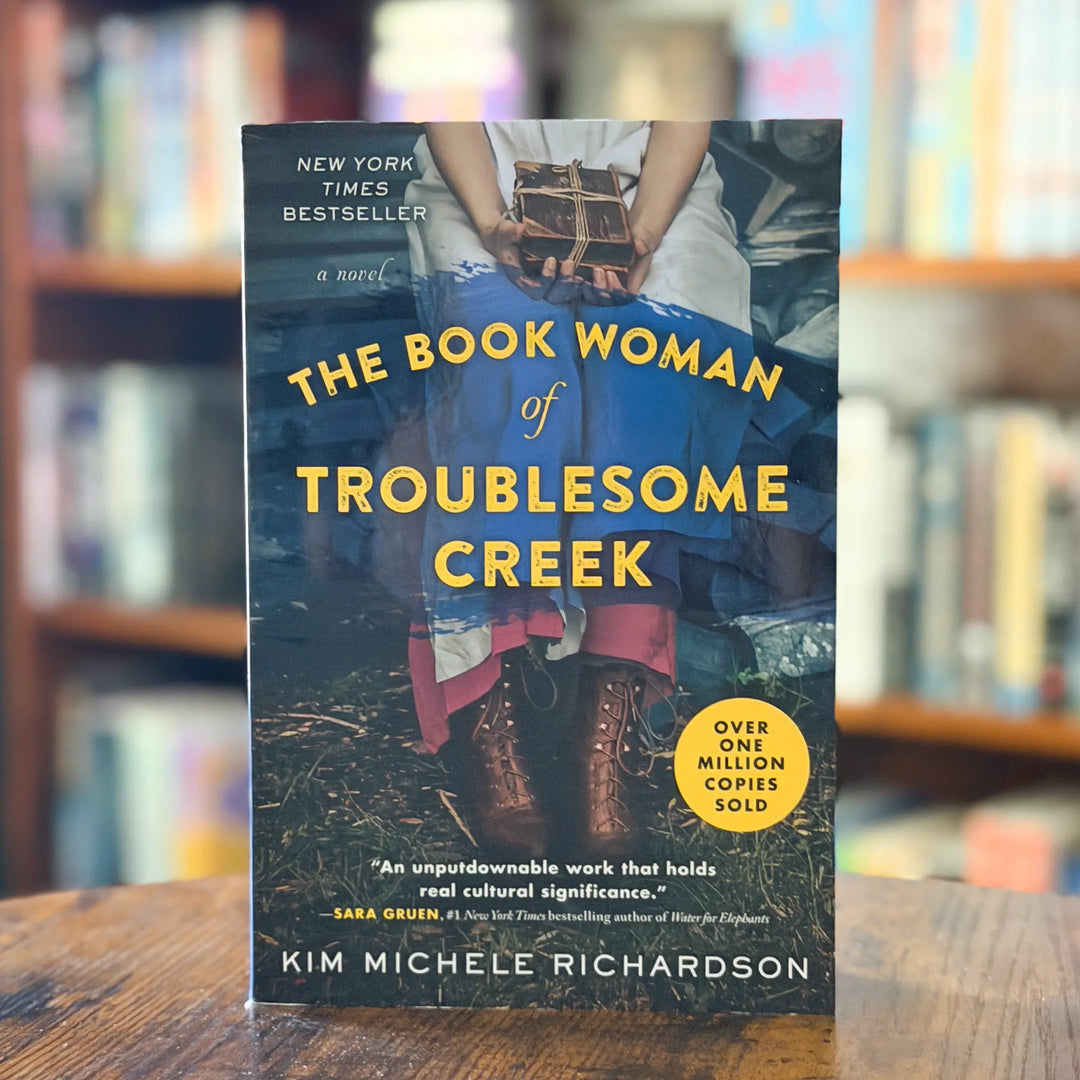 Book cover of 'The Book Woman of Troublesome Creek' by Kim Michele Richardson on a wooden surface with a blurred bookshelf background.