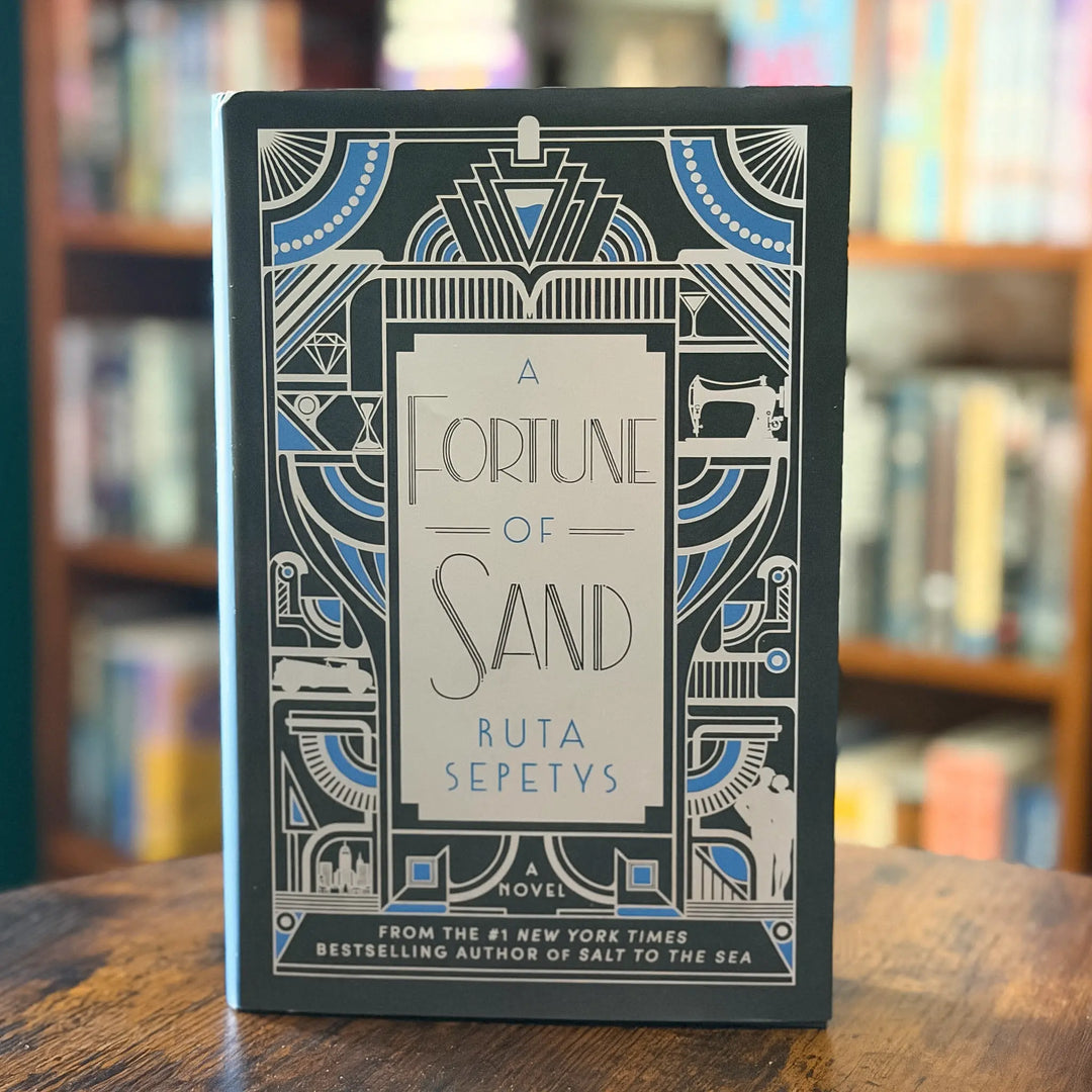Book titled 'A Fortune of Sand' by Ruta Sepetys on a wooden surface with blurred bookshelf background