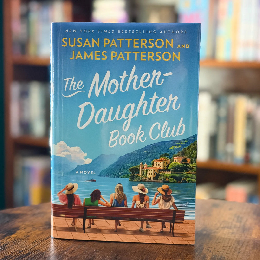 Book cover of 'The Mother-Daughter Book Club' by Susan and James Patterson on a wooden surface with a blurred bookshelf background.