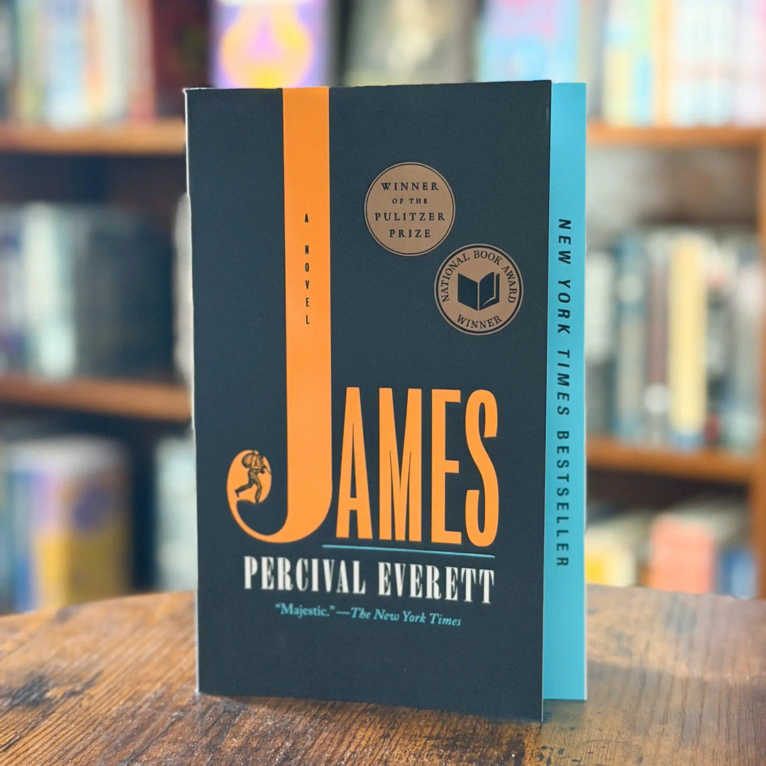 Book titled 'JAMES' by Percival Everett on a wooden surface with a blurred bookshelf background
