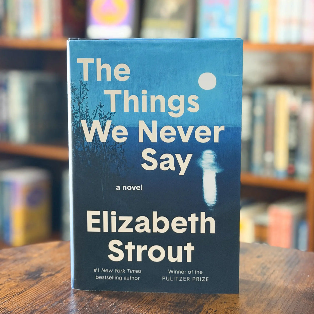Book titled 'The Things We Never Say' by Elizabeth Strout on a wooden surface with a blurred bookshelf background.